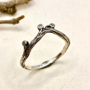 May include: A silver ring with a textured, bark-like finish resembling a tree branch. The band features three small, round, clear stones set in individual bezels at the top, creating a delicate, organic appearance with oxidised detailing.