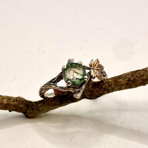 Pode incluir: Um anel com uma pedra de &aacute;gata musgosa verde, redonda e transl&uacute;cida, montada numa banda de prata texturizada que se assemelha a um ramo de &aacute;rvore. O engaste inclui um pequeno detalhe de folha met&aacute;lica que se estende do lado da pedra.