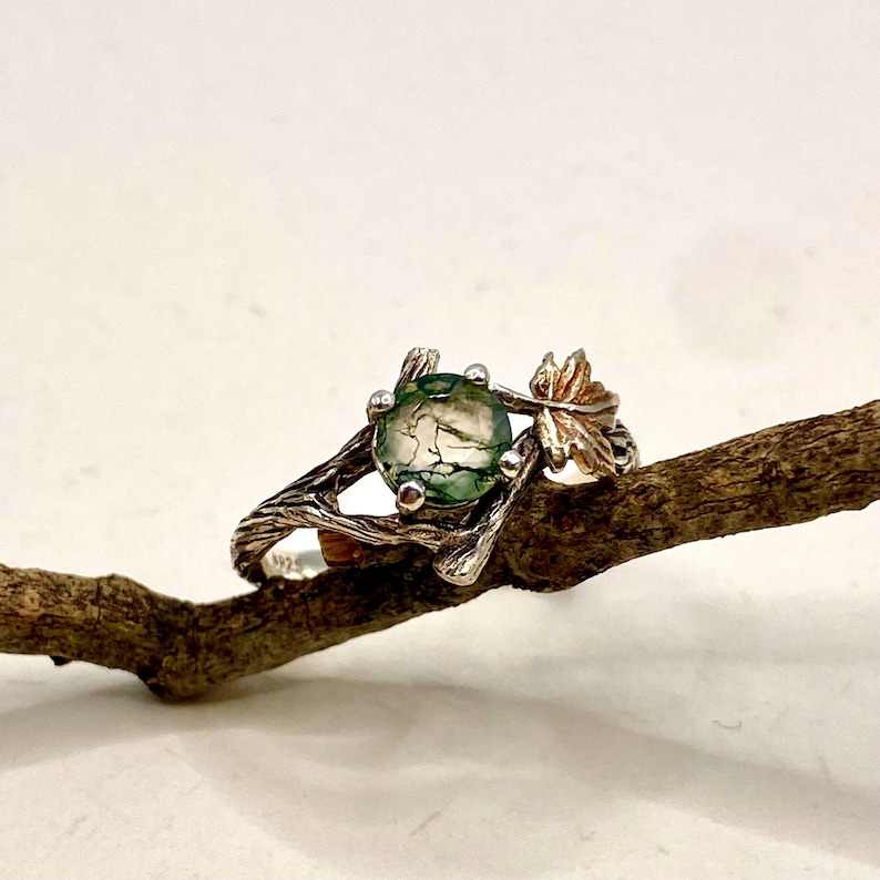 Pode incluir: Um anel com uma pedra de &aacute;gata musgosa verde, redonda e transl&uacute;cida, montada numa banda de prata texturizada que se assemelha a um ramo de &aacute;rvore. O engaste inclui um pequeno detalhe de folha met&aacute;lica que se estende do lado da pedra.