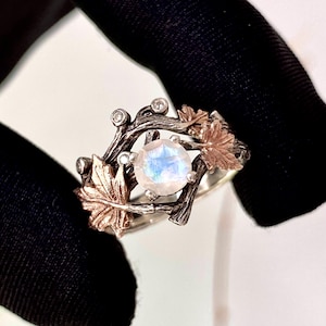 May include: A silver ring with a large, round, iridescent gemstone at the center. The ring features a nature-inspired design with bronze-colored leaves and branches, accented with small, clear gemstones. The ring is set against a dark background.