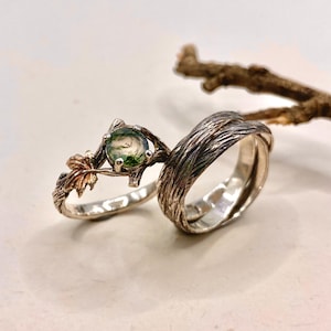 Pode incluir: Um conjunto de dois an&eacute;is composto por uma banda com textura semelhante a casca de &aacute;rvore e um anel com uma pedra redonda de &aacute;gata musgosa verde em crava&ccedil;&atilde;o de garras, decorado com pequenos detalhes de folhas met&aacute;licas numa banda em forma de ramo.