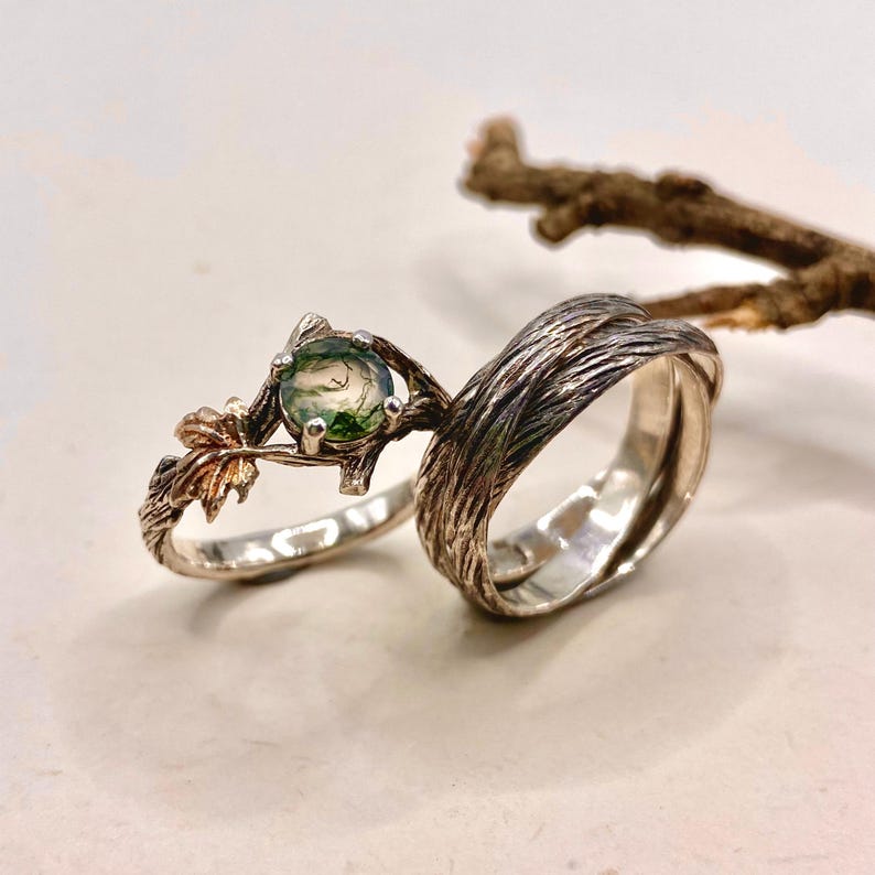 Pode incluir: Um conjunto de dois an&eacute;is composto por uma banda com textura semelhante a casca de &aacute;rvore e um anel com uma pedra redonda de &aacute;gata musgosa verde em crava&ccedil;&atilde;o de garras, decorado com pequenos detalhes de folhas met&aacute;licas numa banda em forma de ramo.