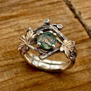 Könnte beinhalten: Ein silberner Ring mit einem von der Natur inspirierten Design. Der Ring hat einen runden, grünen Edelstein in der Mitte, umgeben von einer detaillierten, astähnlichen Fassung mit kleinen, klaren Steinen. Zwei goldfarbene Blätter befinden sich auf beiden Seiten des Edelsteins.