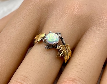 May include: A silver ring with a round, iridescent opal stone. The ring features gold-colored leaf accents on either side of the stone. The ring is designed to look like a leaf and gemstone ring.