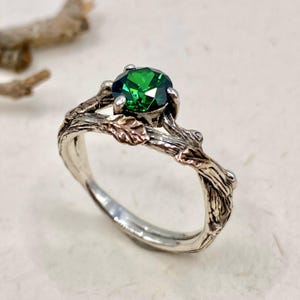 Peut inclure: Une bague en argent ornée d'une pierre précieuse ronde à facettes vert émeraude montée sur un serti à griffes. L'anneau est conçu pour ressembler à une branche d'arbre texturée avec de petites feuilles en relief d'un ton métallique contrasté.