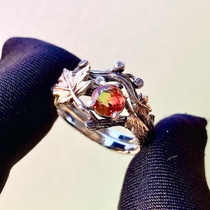 May include: A silver ring with a large, round, multi-colored gemstone. The ring features a nature-inspired design with leaves and branches. Small, clear stones are set around the gemstone. The ring is held against a dark background.