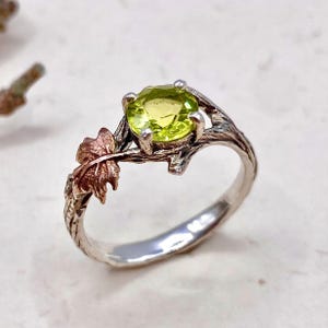Könnte beinhalten: Silberring mit einem hellgrünen, runden Edelstein. Das Ringdesign weist ein Astmotiv mit einem kleinen, kupferfarbenen Blatt auf. Das Band hat ein strukturiertes, organisches Aussehen.