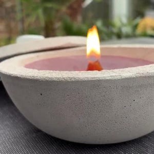 Könnte beinhalten: Ein weißer Beton-Kerzenhalter mit einer brennenden rosa Kerze darin. Der Kerzenhalter hat eine raue, strukturierte Oberfläche.