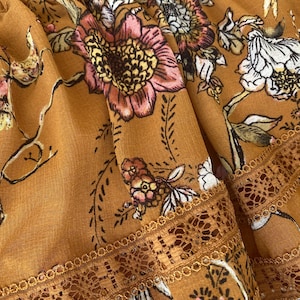 May include: Close-up of a mustard-colored fabric with a floral pattern. The design features pink, white, and yellow flowers with green leaves and brown stems. The fabric has a delicate lace trim along the edge.