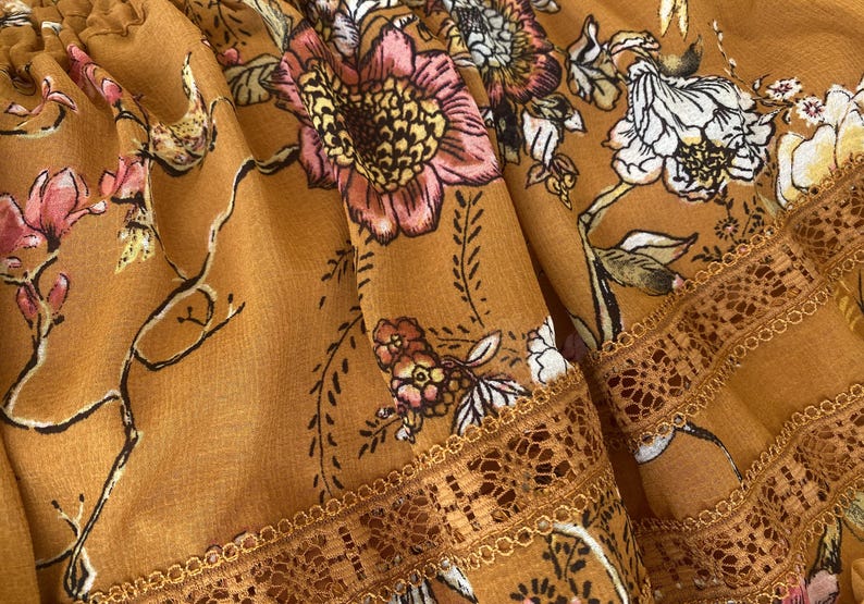May include: Close-up of a mustard-colored fabric with a floral pattern. The design features pink, white, and yellow flowers with green leaves and brown stems. The fabric has a delicate lace trim along the edge.