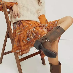 Puede incluir: Conjunto que presenta un suéter crema de manga corta sobre un vestido naranja con estampado floral. Botas de cuero marrón con hebillas completan el look. El conjunto se usa sentado en una silla de madera, creando una estética bohemia.