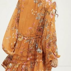 May include: Mustard-colored dress with long, billowy sleeves and a tiered skirt. The dress features a floral pattern in pink, white, and green. The waist is cinched with a drawstring. The dress is made of a lightweight, flowing fabric.