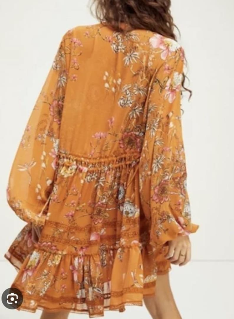 May include: Mustard-colored dress with long, billowy sleeves and a tiered skirt. The dress features a floral pattern in pink, white, and green. The waist is cinched with a drawstring. The dress is made of a lightweight, flowing fabric.