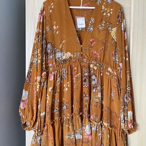 May include: Mustard-colored dress with a floral pattern. The dress has long sleeves, a V-neck, and a tiered skirt. It is hanging on a white hanger against a light-colored door.