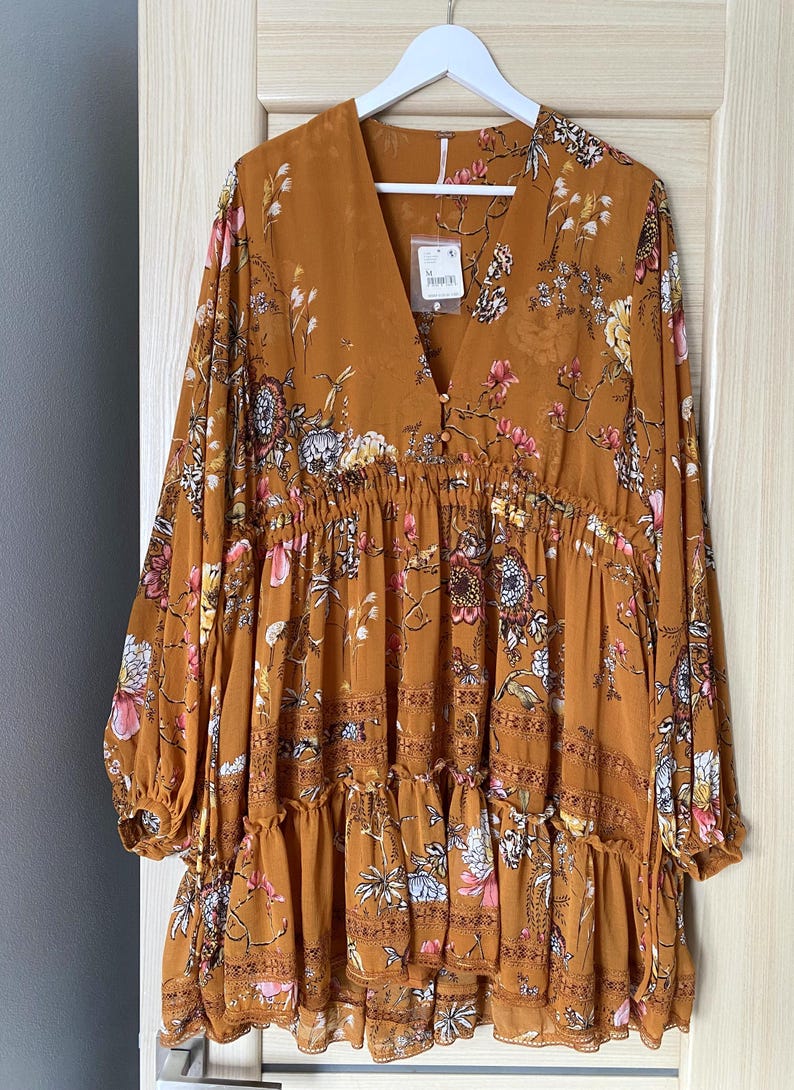 May include: Mustard-colored dress with a floral pattern. The dress has long sleeves, a V-neck, and a tiered skirt. It is hanging on a white hanger against a light-colored door.