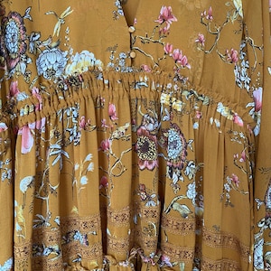 May include: Mustard-colored dress with a V-neck and floral print. The dress features a gathered waist, decorative trim, and a flowing skirt. The floral pattern includes pink, white, and blue flowers with green leaves.