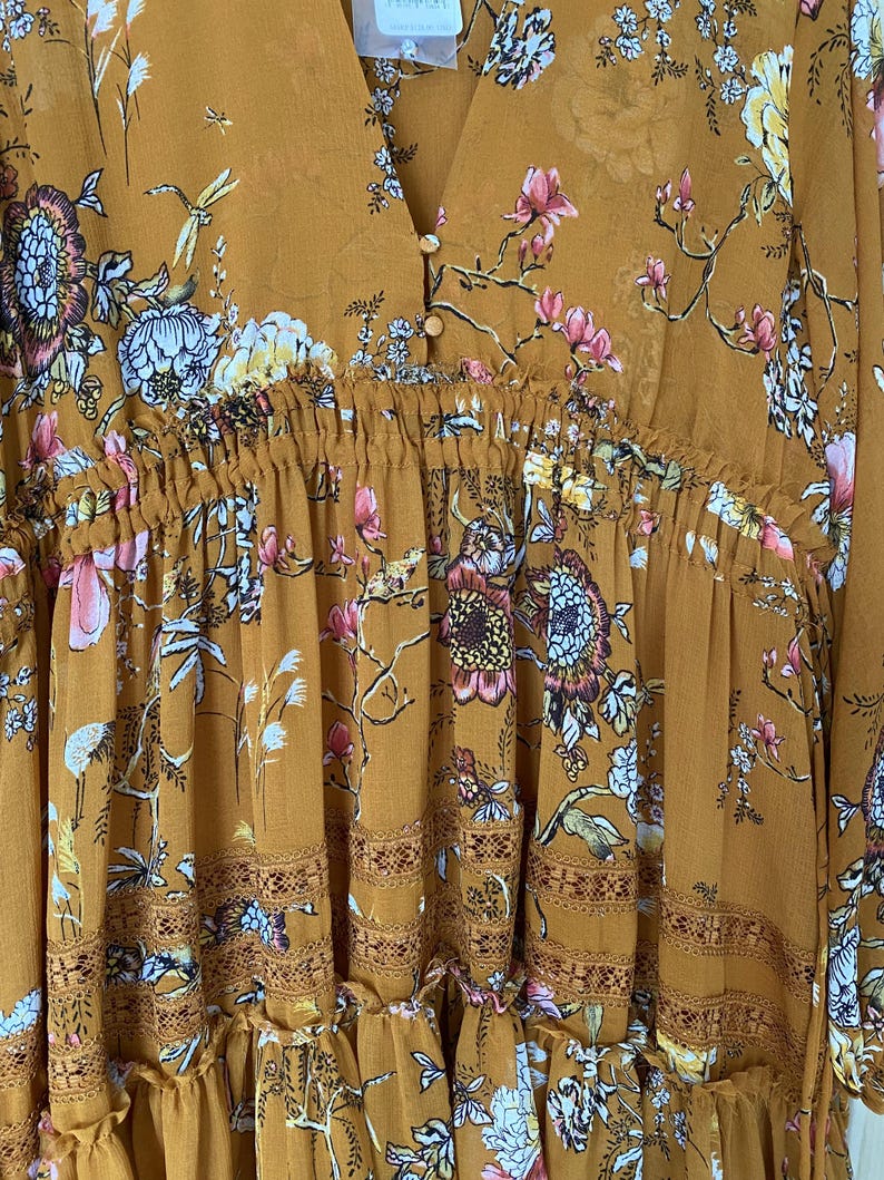 May include: Mustard-colored dress with a V-neck and floral print. The dress features a gathered waist, decorative trim, and a flowing skirt. The floral pattern includes pink, white, and blue flowers with green leaves.