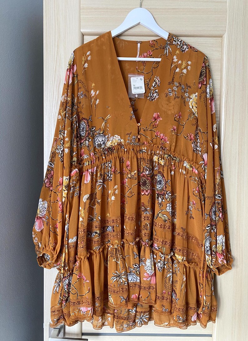 May include: Mustard-colored dress with a floral pattern. The dress has long sleeves, a V-neck, and a tiered skirt. It is hanging on a white hanger against a light-colored wooden door.