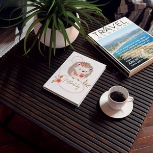 May include: A white notebook with a pink and gold hedgehog illustration and the text "Notebook by" on the cover. The notebook is sitting on a dark wood table with a cup of coffee and a travel magazine.