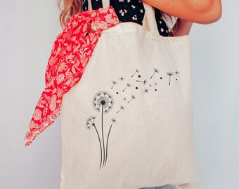 Peut inclure: Un sac fourre-tout beige avec un motif de pissenlit noir. Une écharpe rouge à motifs est visible à l'intérieur du sac. Le sac est tenu par une personne portant un haut à motifs noir et blanc et un jean bleu.