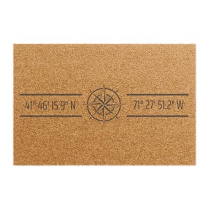 May include: A rectangular doormat made of natural brown coir fiber. The mat features a compass rose design in the center, with the coordinates 41° 46' 15.9" N and 71° 27' 51.2" W printed in dark gray.