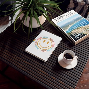 May include: A white journal with the text "NEW YEAR NEW YOU" encircling a smiling face. A "TRAVEL MAGAZINE" and a cup of coffee on a saucer are also on the dark wooden table, along with a potted plant.
