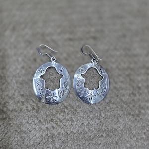 以下が含まれることがあります： シルバーカラーのイヤリングのペア。 各イヤリングは、切り抜かれた手のデザインと、花柄と幾何学模様で囲まれた楕円形を特徴としています。 イヤリングはフック留めです。