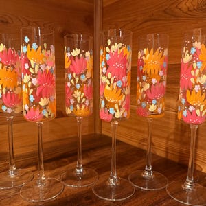 Pu&ograve; includere: Un set di sei fl&ucirc;te da champagne in vetro trasparente con un motivo floreale dipinto a mano in tonalit&agrave; vivaci di arancione, rosa, giallo e azzurro. Ogni bicchiere alto e sottile presenta uno stelo lungo e una base circolare con decori.