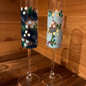 Pu&ograve; includere: Due fl&ucirc;te da champagne in vetro trasparente con disegni floreali dipinti a mano. Il fl&ucirc;te a sinistra presenta fiori blu navy scuro, azzurri e bianchi. Il fl&ucirc;te a destra mostra motivi floreali azzurri, gialli, rosa e verdi sulla superficie.