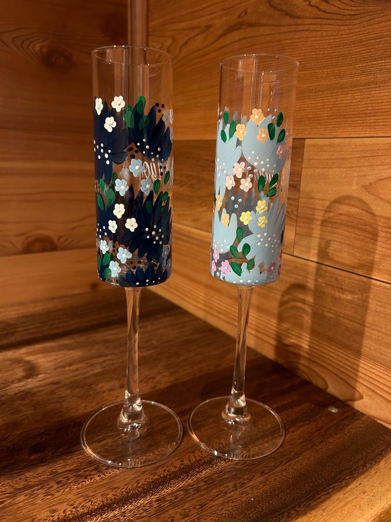 Pu&ograve; includere: Due fl&ucirc;te da champagne in vetro trasparente con disegni floreali dipinti a mano. Il fl&ucirc;te a sinistra presenta fiori blu navy scuro, azzurri e bianchi. Il fl&ucirc;te a destra mostra motivi floreali azzurri, gialli, rosa e verdi sulla superficie.