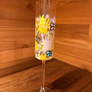 Pu&ograve; includere: Un fl&ucirc;te da champagne in vetro trasparente con un design dipinto a mano di fiori gialli, rosa chiaro e piccoli fiori viola, intervallati da farfalle con contorno nero in blu, giallo e nero, su uno stelo sottile e base rotonda.