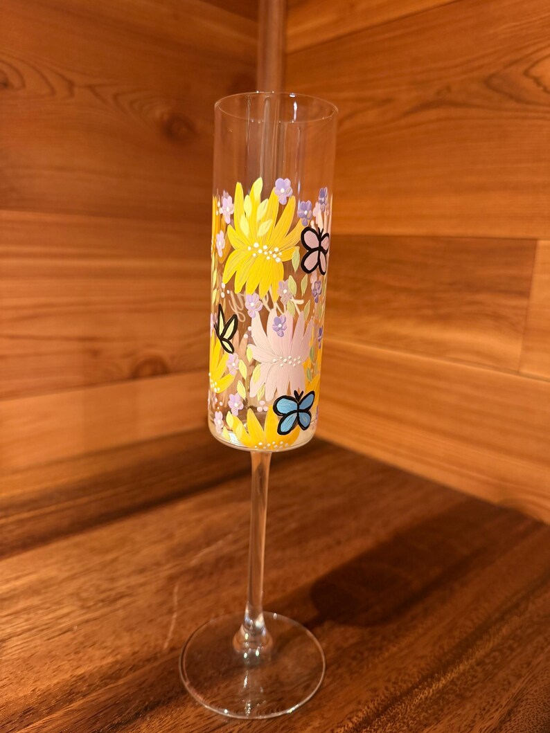 Pu&ograve; includere: Un fl&ucirc;te da champagne in vetro trasparente con un design dipinto a mano di fiori gialli, rosa chiaro e piccoli fiori viola, intervallati da farfalle con contorno nero in blu, giallo e nero, su uno stelo sottile e base rotonda.