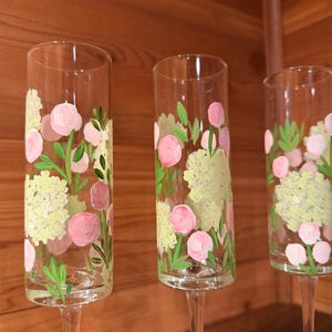 Pu&ograve; includere: Tre fl&ucirc;te da champagne in vetro trasparente con motivi floreali dipinti a mano. Ogni fl&ucirc;te presenta grappoli di fiori giallo pallido simili a ortensie, capolini rosa circolari e sottili foglie verdi dipinte sulla superficie esterna.