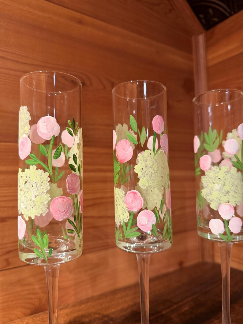 Pu&ograve; includere: Tre fl&ucirc;te da champagne in vetro trasparente con motivi floreali dipinti a mano. Ogni fl&ucirc;te presenta grappoli di fiori giallo pallido simili a ortensie, capolini rosa circolari e sottili foglie verdi dipinte sulla superficie esterna.