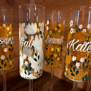 Pu&ograve; includere: Un set di cinque fl&ucirc;te da champagne in vetro trasparente, dipinte a mano con un motivo floreale nei toni del giallo, bianco e verde. Ogni fl&ucirc;te presenta un nome unico in corsivo bianco, tra cui Cassie, Gabby, Susannah, Kate e Isabel.