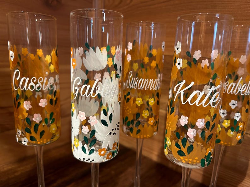 Pu&ograve; includere: Un set di cinque fl&ucirc;te da champagne in vetro trasparente, dipinte a mano con un motivo floreale nei toni del giallo, bianco e verde. Ogni fl&ucirc;te presenta un nome unico in corsivo bianco, tra cui Cassie, Gabby, Susannah, Kate e Isabel.