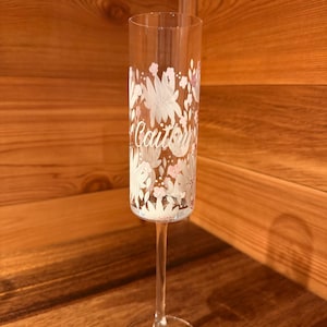 Pu&ograve; includere: Un fl&ucirc;te da champagne in vetro trasparente con un motivo floreale bianco e rosa chiaro dipinto sull'esterno. Il nome Caitlyn &egrave; scritto in corsivo bianco al centro del disegno floreale sulla superficie del vetro.