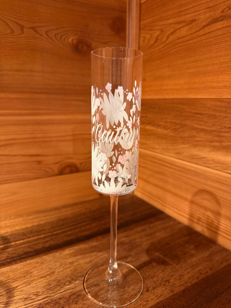 Pu&ograve; includere: Un fl&ucirc;te da champagne in vetro trasparente con un motivo floreale bianco e rosa chiaro dipinto sull'esterno. Il nome Caitlyn &egrave; scritto in corsivo bianco al centro del disegno floreale sulla superficie del vetro.
