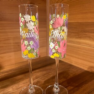 Pu&ograve; includere: Due fl&ucirc;te da champagne in vetro trasparente con motivi floreali dipinti a mano nei toni del rosa, giallo, bianco e viola. Un calice &egrave; personalizzato con il nome Hailey in corsivo bianco, l'altro con il nome Ethan in corsivo bianco.