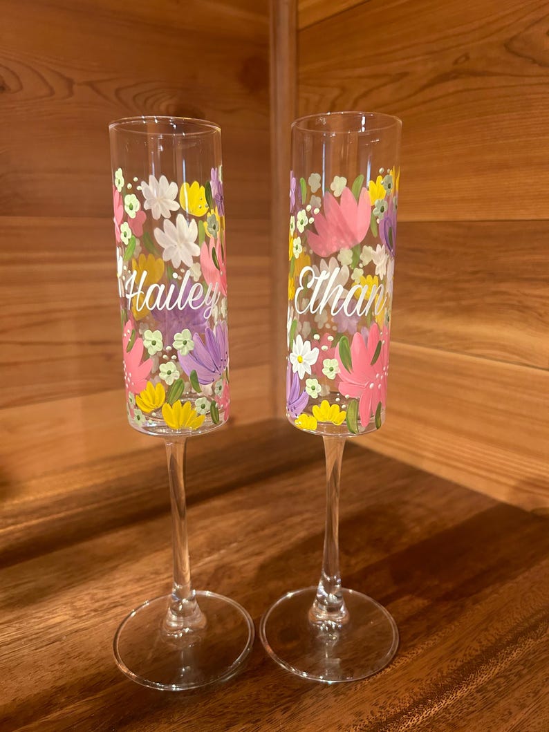 Pu&ograve; includere: Due fl&ucirc;te da champagne in vetro trasparente con motivi floreali dipinti a mano nei toni del rosa, giallo, bianco e viola. Un calice &egrave; personalizzato con il nome Hailey in corsivo bianco, l'altro con il nome Ethan in corsivo bianco.