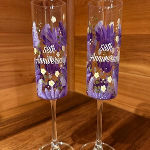 Pu&ograve; includere: Due fl&ucirc;te da champagne in vetro trasparente con motivi floreali viola dipinti a mano e la scritta bianca 58th Anniversary. Ogni fl&ucirc;te presenta un corpo cilindrico alto e sottile, uno stelo sottile e una base circolare con petali viola.