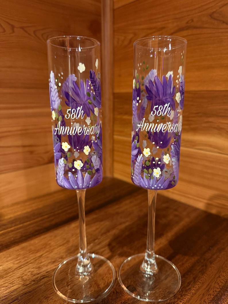 Pu&ograve; includere: Due fl&ucirc;te da champagne in vetro trasparente con motivi floreali viola dipinti a mano e la scritta bianca 58th Anniversary. Ogni fl&ucirc;te presenta un corpo cilindrico alto e sottile, uno stelo sottile e una base circolare con petali viola.