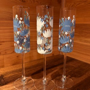 Pu&ograve; includere: Tre fl&ucirc;te da champagne in vetro trasparente con steli alti e sottili e basi circolari. Le superfici in vetro sono dipinte a mano con un motivo floreale caratterizzato da petali blu e bianchi, piccoli punti bianchi e foglie verdi in un design ripetuto.