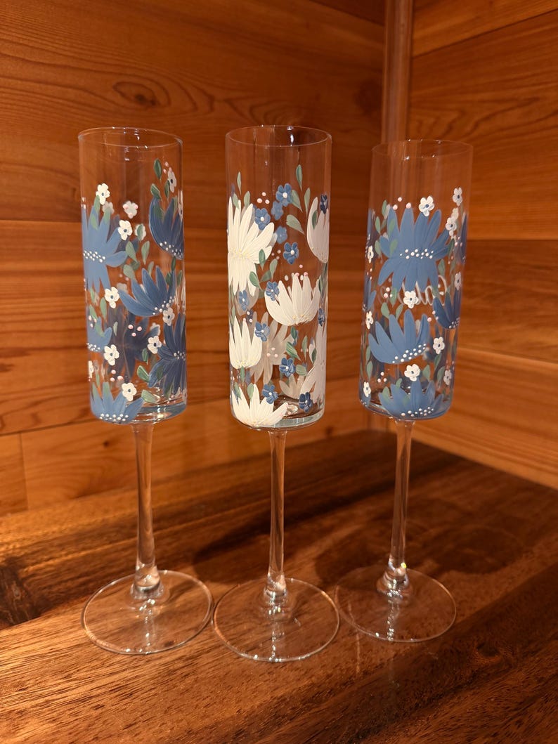 Pu&ograve; includere: Tre fl&ucirc;te da champagne in vetro trasparente con steli alti e sottili e basi circolari. Le superfici in vetro sono dipinte a mano con un motivo floreale caratterizzato da petali blu e bianchi, piccoli punti bianchi e foglie verdi in un design ripetuto.