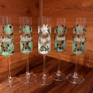 Pu&ograve; includere: Cinque fl&ucirc;te da champagne in vetro trasparente con un motivo dipinto a mano di foglie verde menta e piccoli fiori neri. Ogni fl&ucirc;te &egrave; personalizzato con un nome in corsivo bianco: Ashley, Christina, Payton, Jenna e Andrea, disposti in fila.