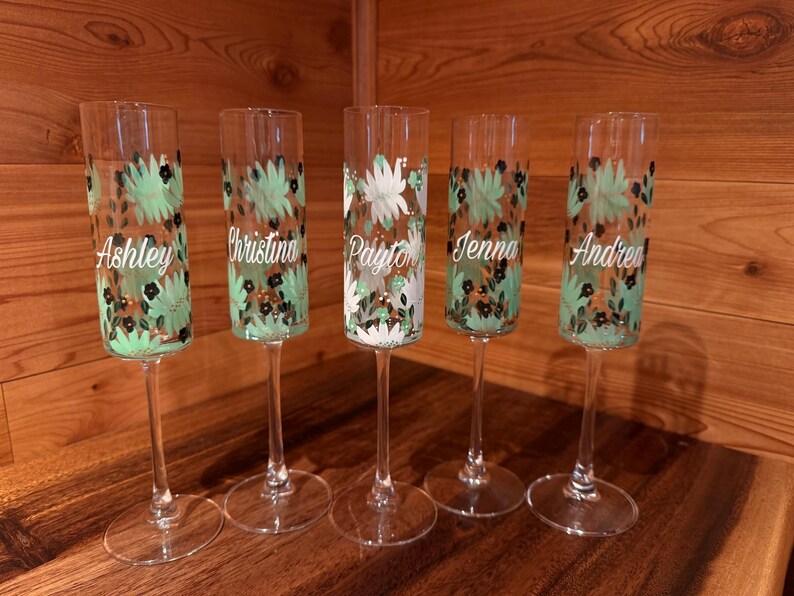 Pu&ograve; includere: Cinque fl&ucirc;te da champagne in vetro trasparente con un motivo dipinto a mano di foglie verde menta e piccoli fiori neri. Ogni fl&ucirc;te &egrave; personalizzato con un nome in corsivo bianco: Ashley, Christina, Payton, Jenna e Andrea, disposti in fila.