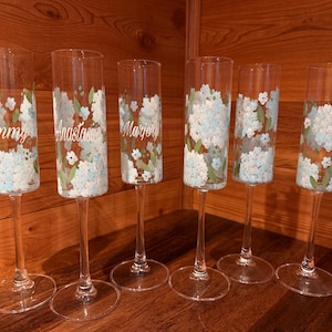 Pu&ograve; includere: Sei fl&ucirc;te da champagne in vetro trasparente con grappoli di fiori dipinti a mano in bianco e azzurro con foglie verdi. Tre dei calici sono personalizzati con i nomi Tammy, Anastasia e Marjory stampati in corsivo bianco sul vetro.