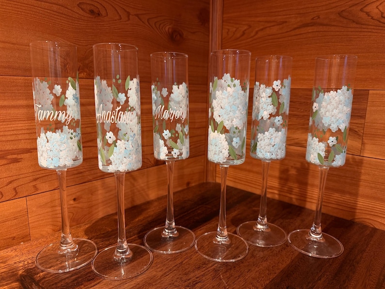 Pu&ograve; includere: Sei fl&ucirc;te da champagne in vetro trasparente con grappoli di fiori dipinti a mano in bianco e azzurro con foglie verdi. Tre dei calici sono personalizzati con i nomi Tammy, Anastasia e Marjory stampati in corsivo bianco sul vetro.