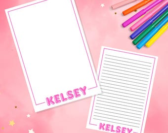 May include: Two white rectangular notepads with pink borders and the name KELSEY printed in pink bubble letters at the bottom. One notepad is blank, and the other features horizontal black lines. Six colorful felt-tip pens are arranged nearby.