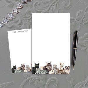 May include: White notepad and stationery set featuring a watercolor illustration of various cat breeds at the bottom. The notepad has the text "ADD A NAME OR TEXT!". A black and gold pen is also included.